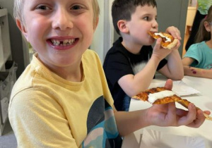 Chłopiec uśmiecha się do zdjęcia. W ręku trzyma kawałek pizzy. Drugi chłopiec w tle je pizzę